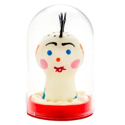 Spielzeugfigur mit Gesicht, bunten Details und schwarzem Haar, in einer durchsichtigen Kuppel auf rotem Sockel.